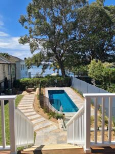 View from a porch overlooking a small rectangular swimming pool, surrounded by a fence and garden, with large trees, a stone path, and a glimpse of water in the background under a blue sky.