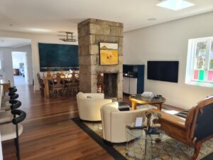 Bright, modern living and dining area with wooden floors, stone divider, and contemporary furniture. Large dining table in back, white armchairs, TV, colorful art, and light streaming through a window on the right.
