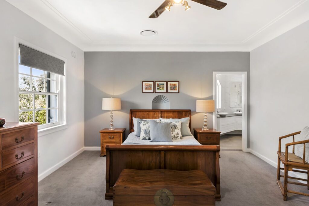 A bright bedroom with a wooden bed, two bedside tables with lamps, a wooden chest at the foot of the bed, gray carpet, a window with a blind, a wooden chair, and an open door leading to a bathroom.