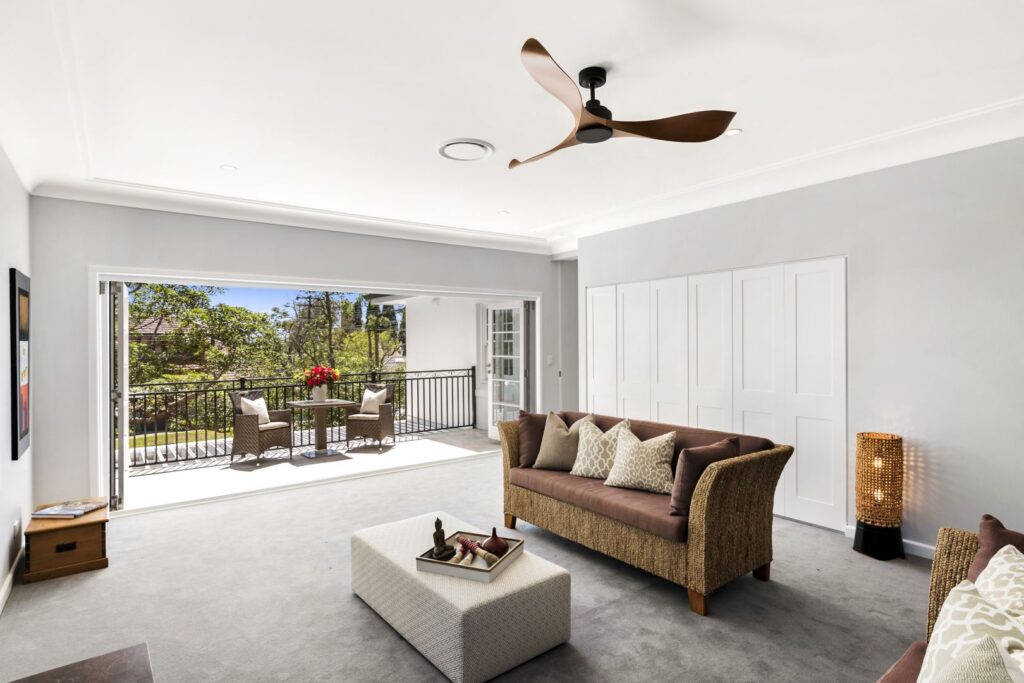 Spacious living room with a brown sofa, ottoman, and neutral decor. Large sliding doors open to a balcony with outdoor seating and greenery visible outside. A ceiling fan hangs above.