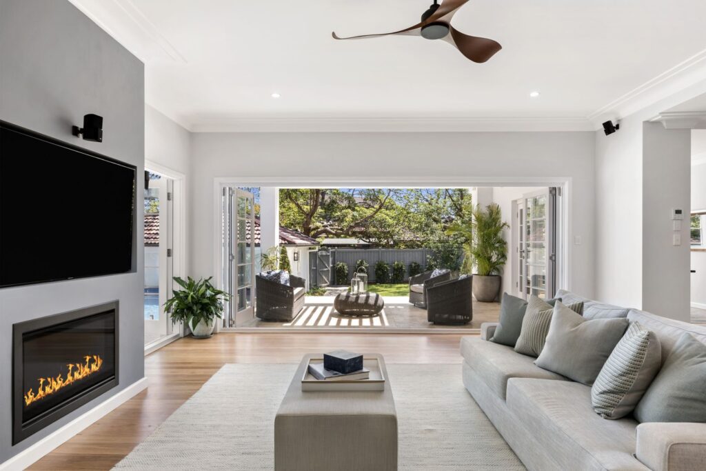 Modern living room with a gray sofa, wall-mounted TV above a fireplace, houseplants, and glass doors opening to a sunny patio with outdoor chairs and greenery. Natural light fills the space.