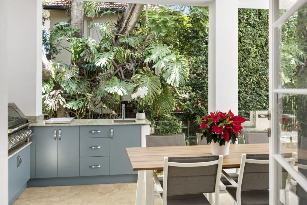 Modern outdoor kitchen with blue cabinets, a built-in grill, and a wooden dining table set with gray chairs. A potted red poinsettia decorates the table. Lush greenery and trees surround the patio area.