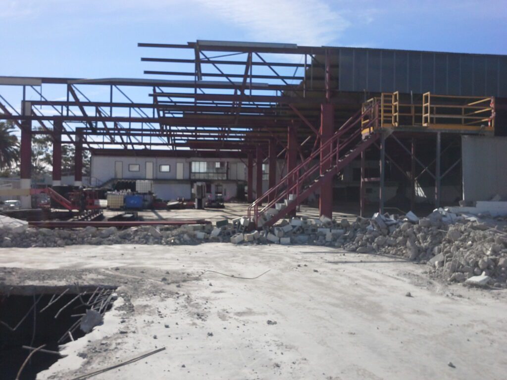 A partially constructed or demolished industrial building with exposed steel beams, rubble on the ground, and a red metal staircase leading to an upper level under a clear sky.
