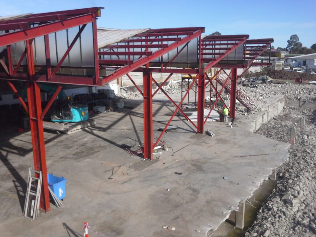 Partially constructed steel framework of a building on a concrete foundation, with construction equipment and rubble nearby. Two workers are visible near the center, and the sky is clear.