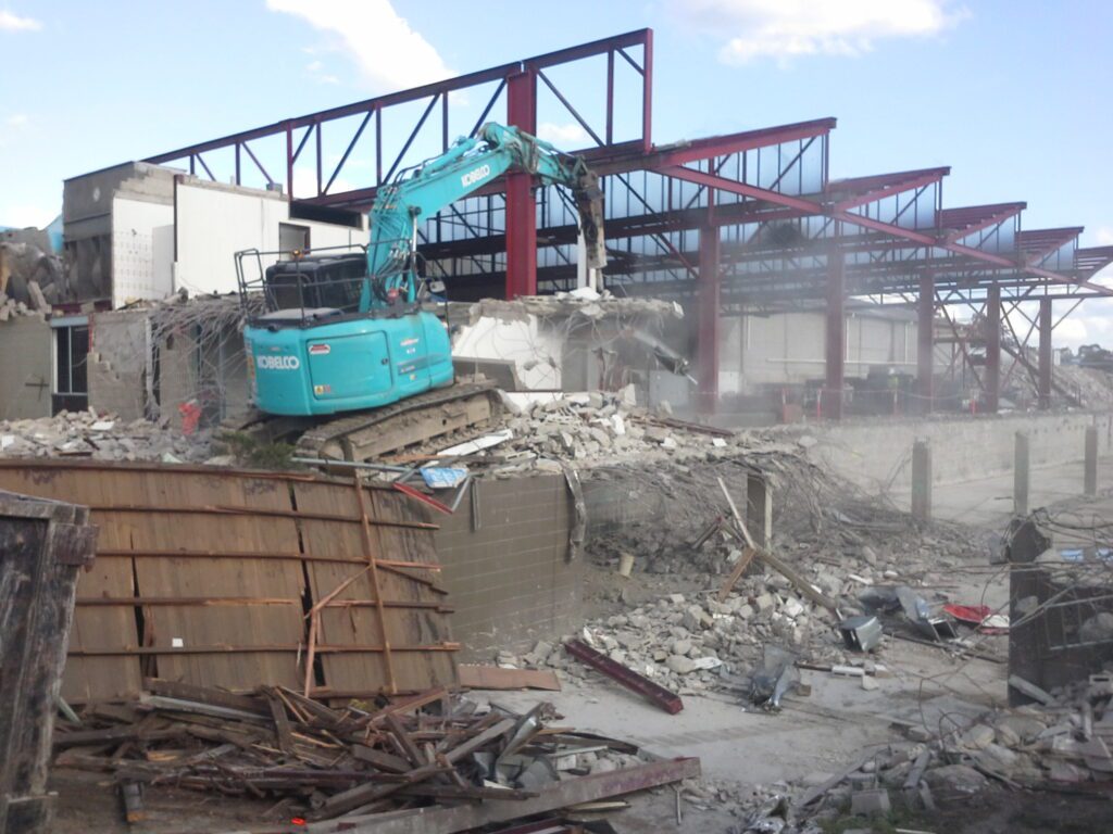 A blue excavator demolishes a concrete building, with debris and rubble scattered around. In the background, a steel framework of a large unfinished structure is visible under a partly cloudy sky.
