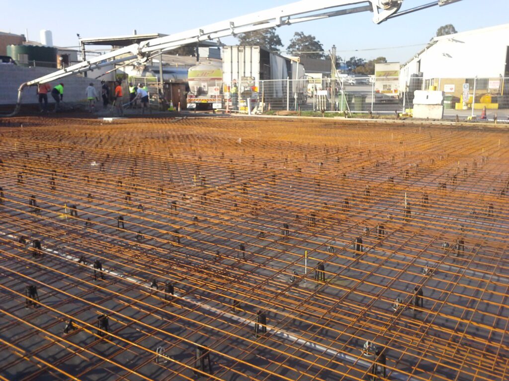 A construction site shows a large area covered with a grid of steel rebar, prepared for concrete pouring. Construction equipment and workers are visible in the background.
