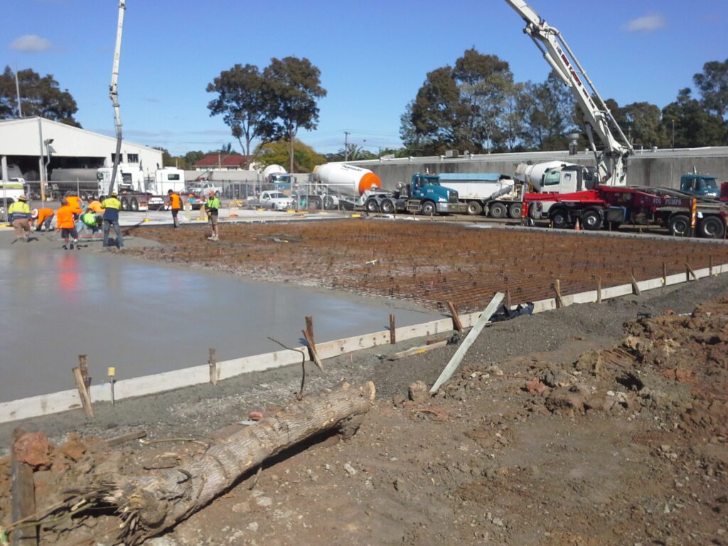 Construction workers pour and level concrete on a building site. Several concrete mixer trucks and machinery are present, with rebar exposed and trees in the background under a clear sky.