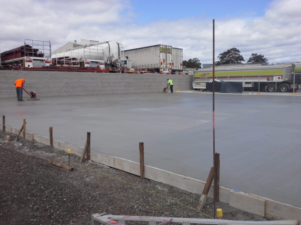 Construction workers in high-visibility clothing smooth a large, freshly poured concrete slab at an outdoor construction site, with trucks and equipment in the background under a cloudy sky.