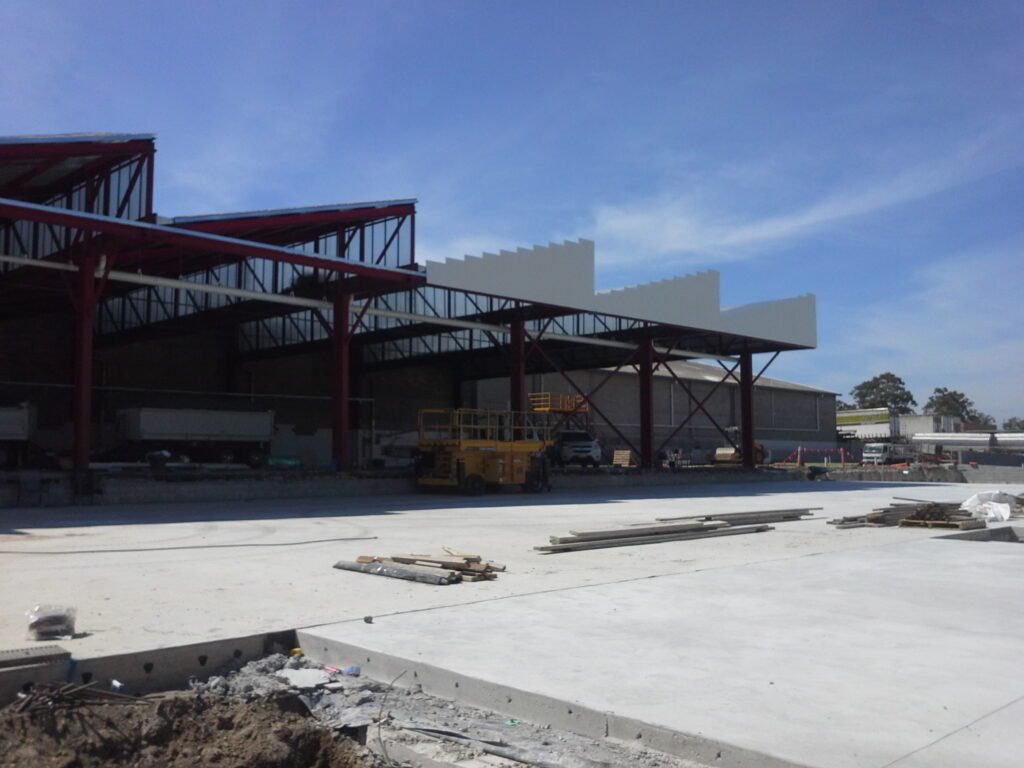 Partially constructed industrial building with red steel beams, some roof panels installed, and construction equipment and materials scattered on a large concrete slab under a blue sky.