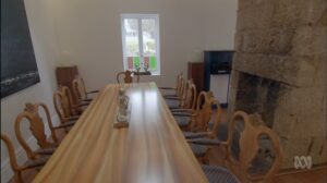 A long wooden dining table with ten ornate chairs is set in a bright room with white walls, a stone fireplace, and a window letting in natural light. Small decorative items are placed on the table.