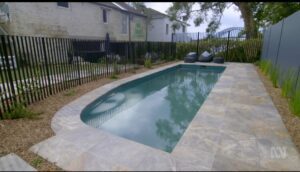 A backyard swimming pool with gray stone tiles, surrounded by a black metal fence, landscaping rocks, and two black lounge chairs at the far end near trees and neighboring houses.