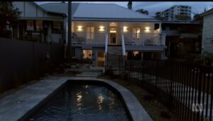 A modern house with bright lights is seen at dusk, featuring a balcony, a backyard swimming pool, stone paving, and a fenced garden. Apartment buildings are visible in the background under a cloudy evening sky.