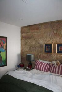 A cozy bedroom with a stone accent wall, two striped pillows, a teddy bear on the bed, a lamp on a bedside table, and colorful artwork hanging on the walls.