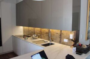 Modern kitchen with gray cabinets, marble countertops, a sink, and a built-in stovetop. Under-cabinet lighting highlights a stone backsplash. A laptop and other small items are visible on the counter.
