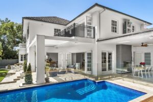 Modern two-story white house with large windows, a balcony, and an outdoor dining area. There is a swimming pool with glass fencing, poolside plants, and a shaded patio area, all under a clear blue sky.