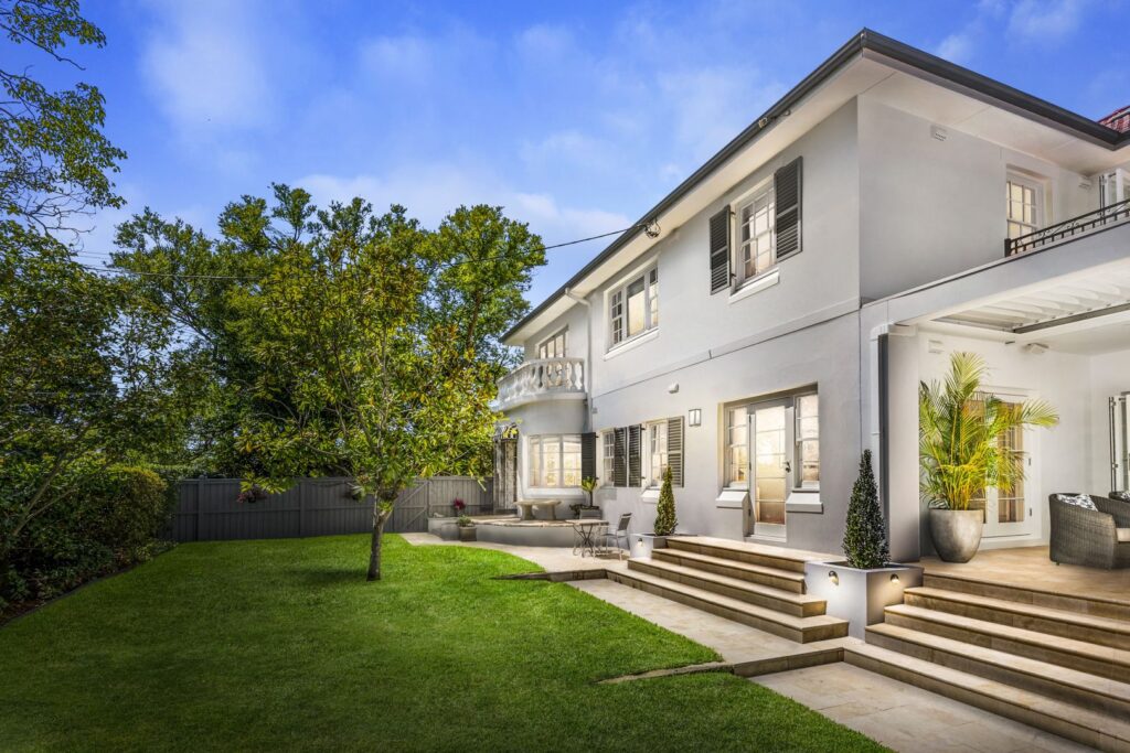 A modern two-story white house with large windows and a balcony, surrounded by a lush green lawn, garden, and trees. The patio area features seating and outdoor lighting, and steps lead up to the main entrance.
