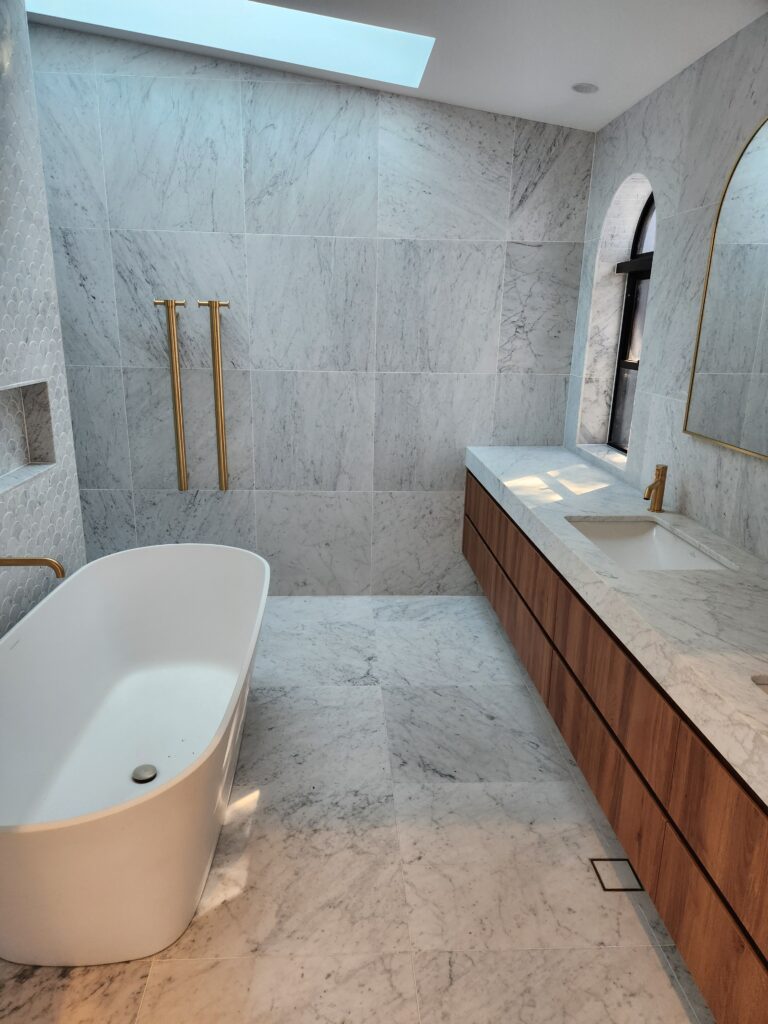 Modern bathroom with marble walls and floors, a white freestanding bathtub, gold fixtures, wooden vanity with twin sinks, and a skylight and arched window allowing natural light.