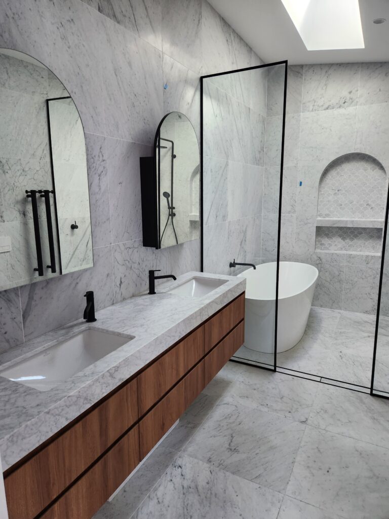 Modern bathroom with marble walls and floors, a floating wooden vanity with two sinks, black fixtures, a freestanding white bathtub, and a glass shower partition. Large mirrors hang above the vanity.