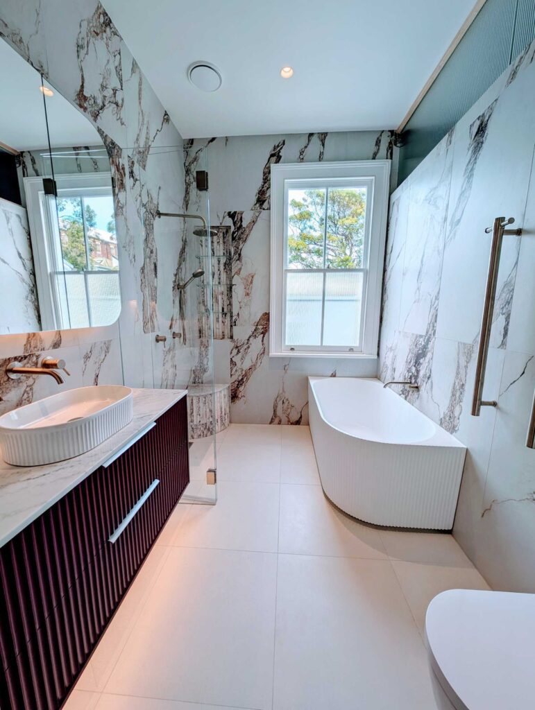 Modern bathroom with marble walls, a freestanding white bathtub by a window, a large mirror above a vanity with a vessel sink, and a glass-enclosed shower. Natural light enters through the window.
