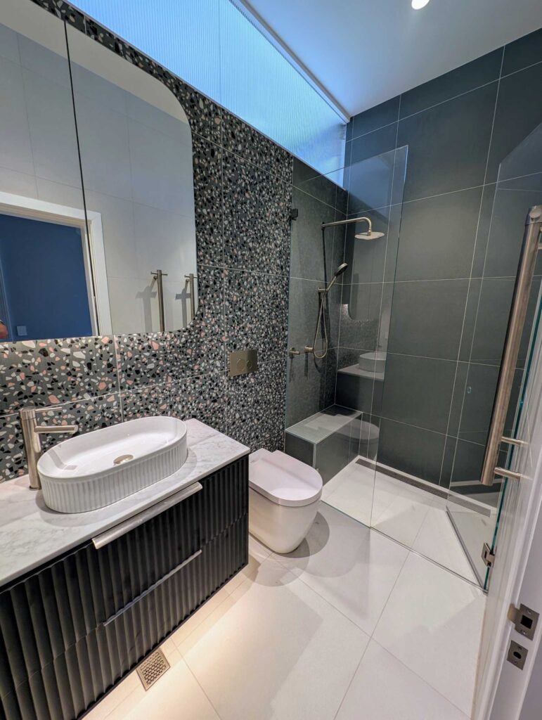 Modern bathroom with terrazzo accent wall, vessel sink on a dark vanity, wall-mounted toilet, and a glass-enclosed shower with a rainfall showerhead. The room features sleek, minimalist design and neutral colors.
