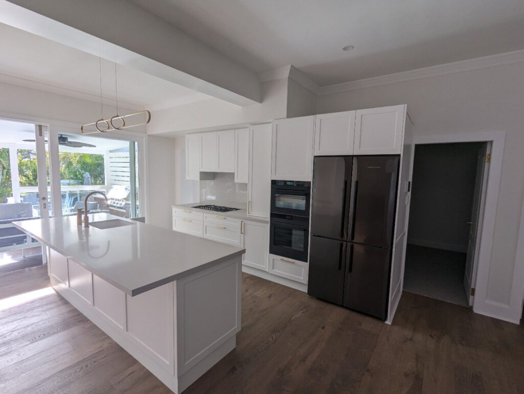Modern kitchen with white cabinets, a large island with a sink, built-in gas cooktop, double oven, and a dark refrigerator. Large windows and glass doors let in natural light and provide an outdoor view.