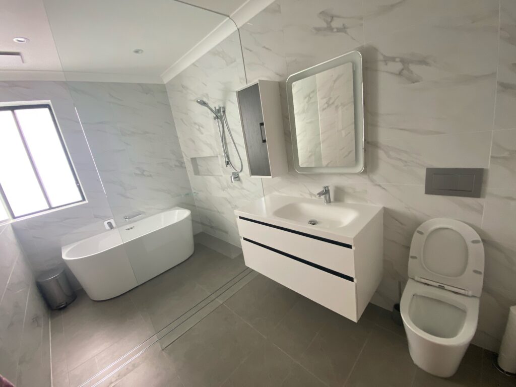 Modern bathroom with a white bathtub, wall-mounted shower, floating vanity with sink and mirror, and a toilet. The walls have marble-style tiles and there’s a window letting in natural light.