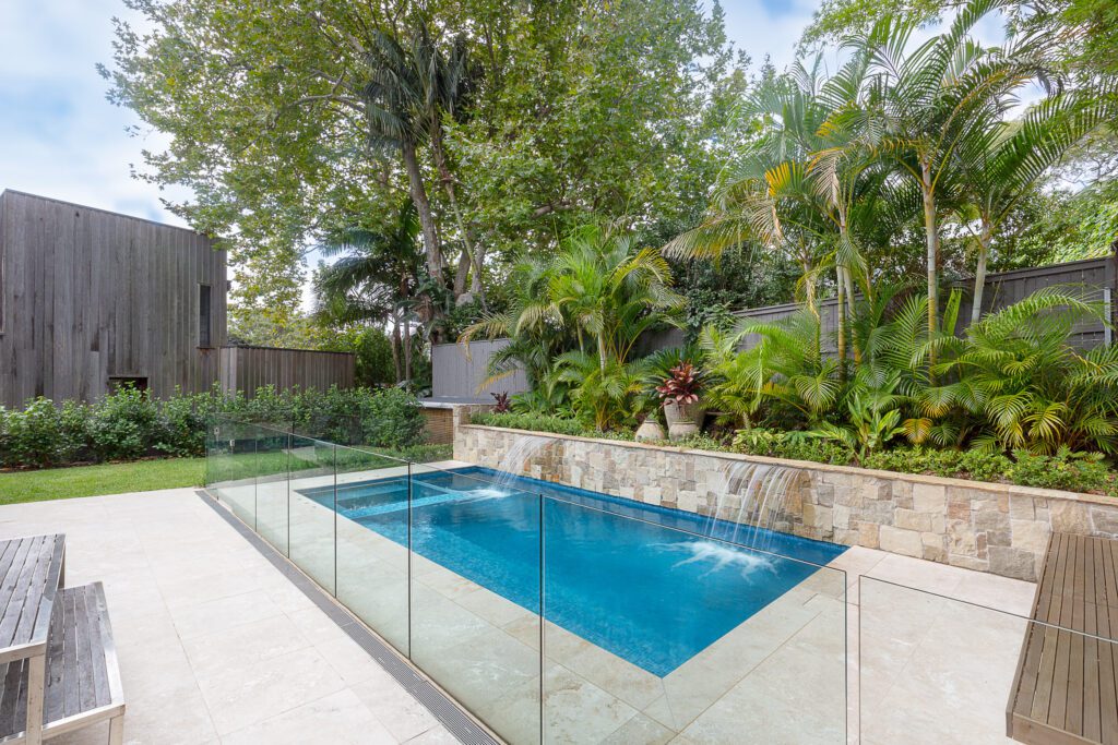Modern backyard pool with stone-tiled edge, glass safety fence, water feature, and surrounding tropical plants; a wooden bench and building are visible to the left, with tall trees in the background.