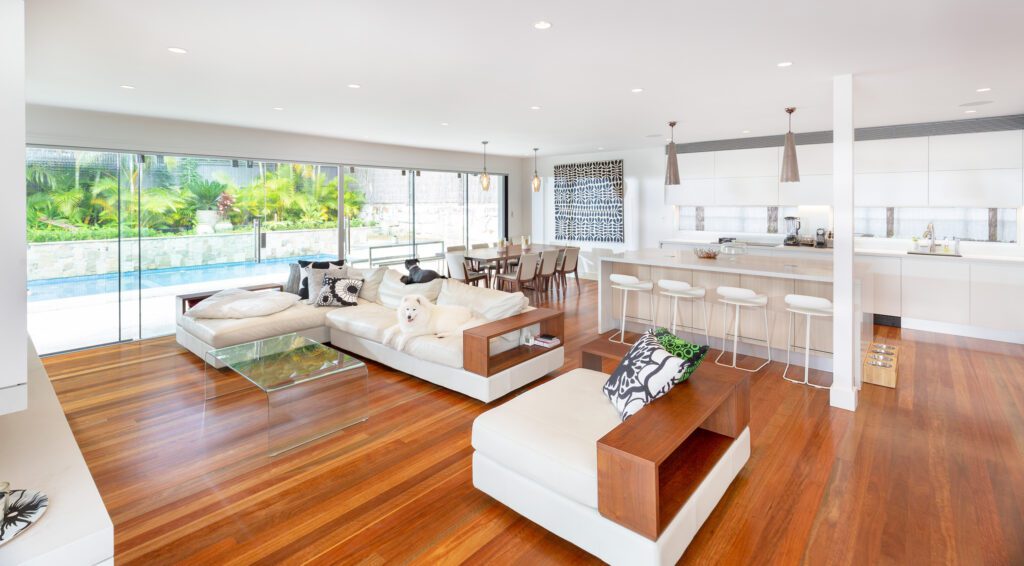 Spacious, modern open-plan living area with wood floors, white sofas, a kitchen with bar stools, dining table, and large glass doors overlooking a pool and lush greenery outside.
