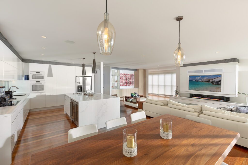Modern open-plan kitchen and living area with white cabinetry, marble island, wooden dining table, pendant lights, beige sectional sofa, and wall-mounted TV displaying a scenic beach view. Natural light fills the space.