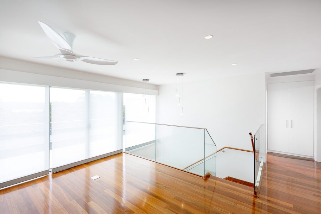 A bright, modern interior with polished wooden floors, white walls, a ceiling fan, glass stair railing, pendant lights, and large windows with sheer blinds letting in natural light.