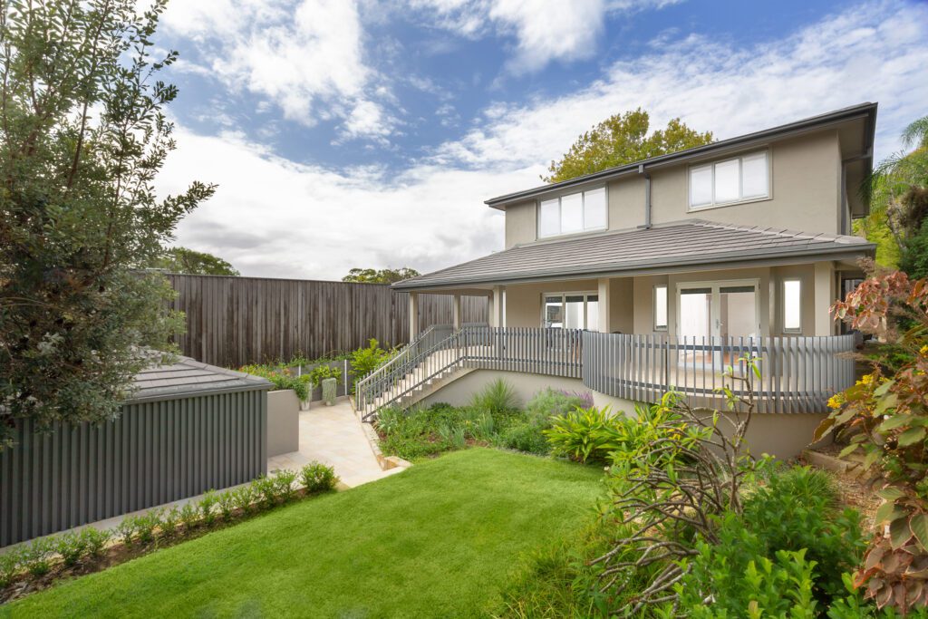 Modern two-story house with beige exterior, large windows, and a wraparound balcony. The yard features well-kept grass, lush plants, a small shed, and is enclosed by a wooden fence under a partly cloudy sky.