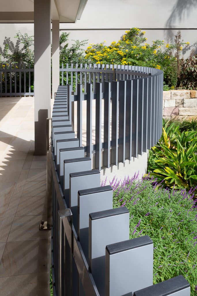 A modern garden features a curved metal fence with rectangular vertical posts, surrounded by lush greenery and colorful flowers, including purple and yellow blooms. Sunlight casts shadows on the patio area.