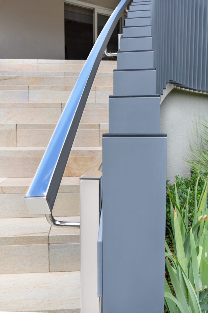 Close-up view of a modern staircase with light stone steps and a sleek metal handrail attached to blue-gray square posts, with green plants visible on the right side.