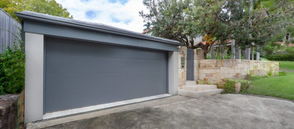 A modern gray garage door with smooth panels, surrounded by stone walls and a paved driveway. Steps lead up to a gate beside the garage, with trees and green lawn in the background.
