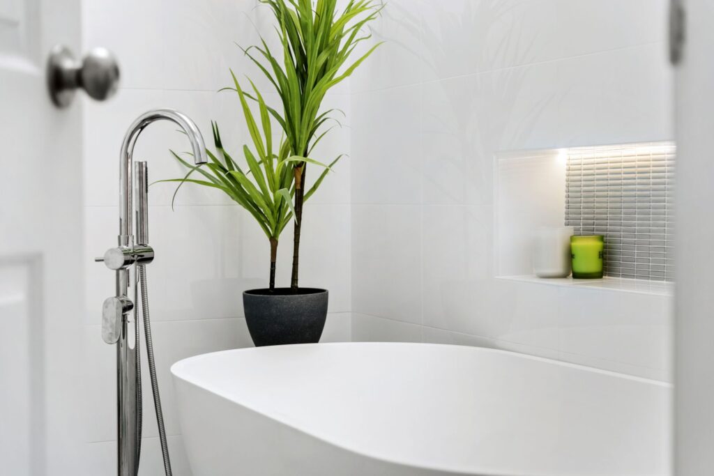 A modern white bathroom with a freestanding bathtub, a tall green potted plant, and a recessed shelf holding candles and a green container, all against white tiled walls.