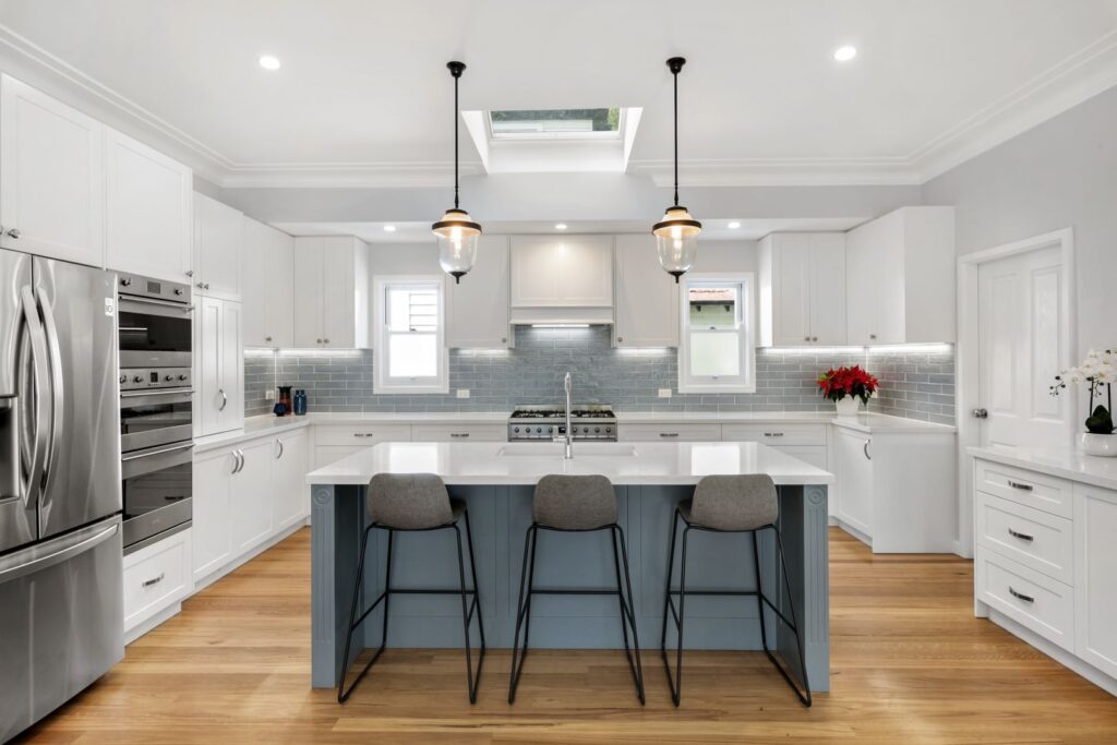 Modern kitchen with white cabinets, stainless steel appliances, a blue island with three bar stools, pendant lights, light wood flooring, and a skylight above. A vase of red flowers and an orchid add color to the space.