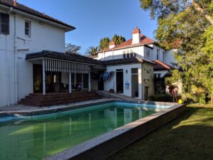 A large white house with red roof tiles sits beside a rectangular swimming pool with green water. Laundry hangs on a line near a covered porch, and trees provide shade in the garden.