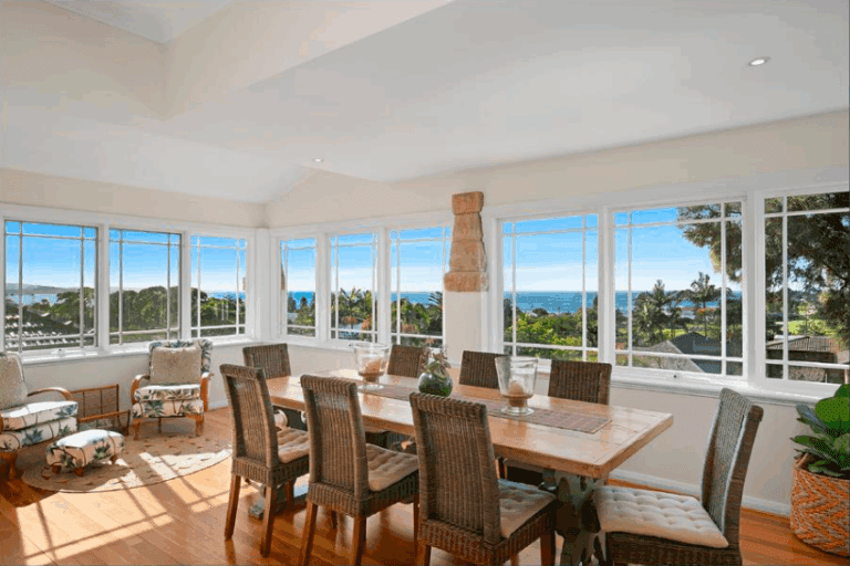 Bright dining room with large windows, wicker chairs around a wooden table, potted plants, and ocean views with blue sky and palm trees visible outside.