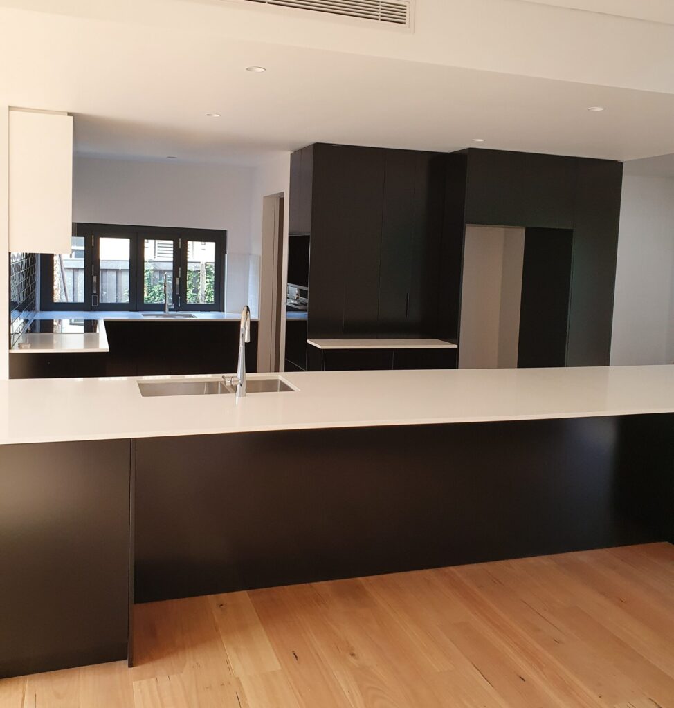 A modern kitchen with black cabinets, white countertops, a large island with a sink, light wood flooring, and large windows letting in natural light.