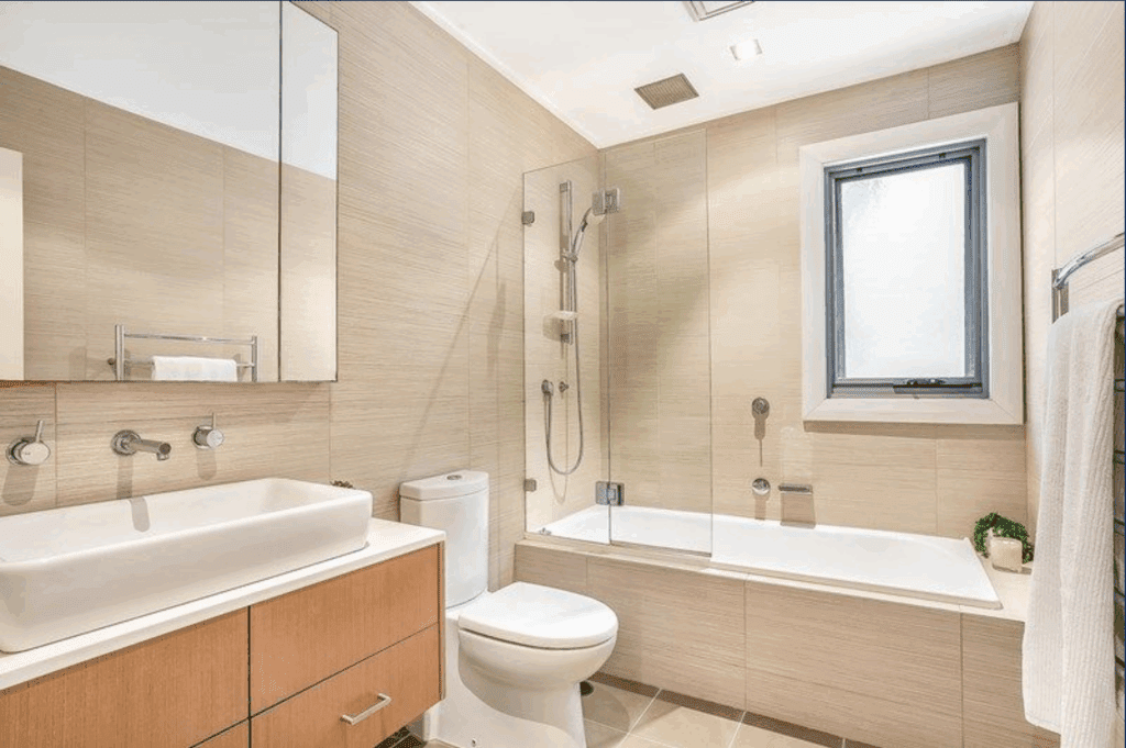 Modern bathroom with beige tile walls, a floating wood vanity with vessel sink, wall-mounted faucet, toilet, and a bathtub with glass shower screen. There is a window, towel rack, and small green plants for decor.