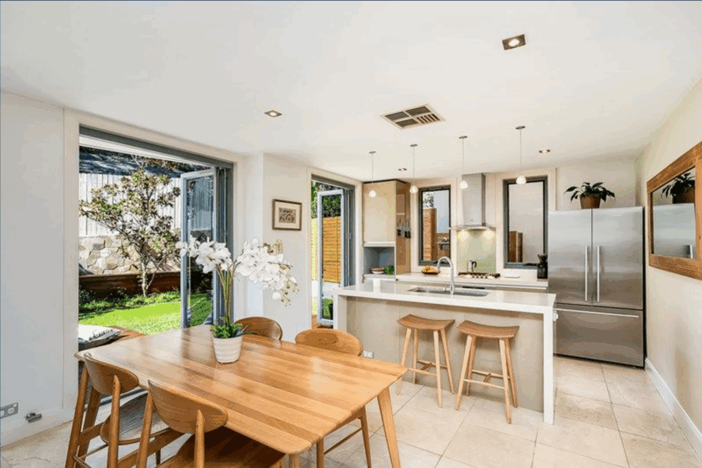 Modern kitchen and dining area with wooden furniture, white countertops, stainless steel appliances, and large windows opening to a green backyard. An orchid centerpiece is on the dining table. The space is bright and airy.