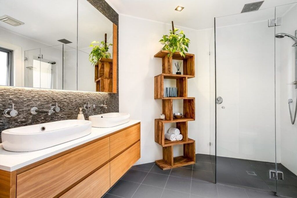 Modern bathroom with double sinks, wooden vanity, and a tall wooden shelf holding towels and decor. A glass-enclosed shower is on the right, and green plants add a fresh touch. The space is bright and contemporary.