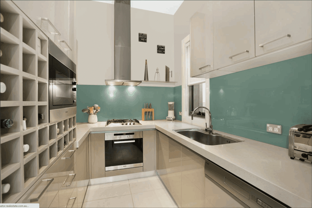 Modern kitchen with glossy beige cabinets, a built-in oven, gas stovetop, wine rack, stainless steel appliances, turquoise backsplash, and under-cabinet lighting. A window above the sink lets in natural light.