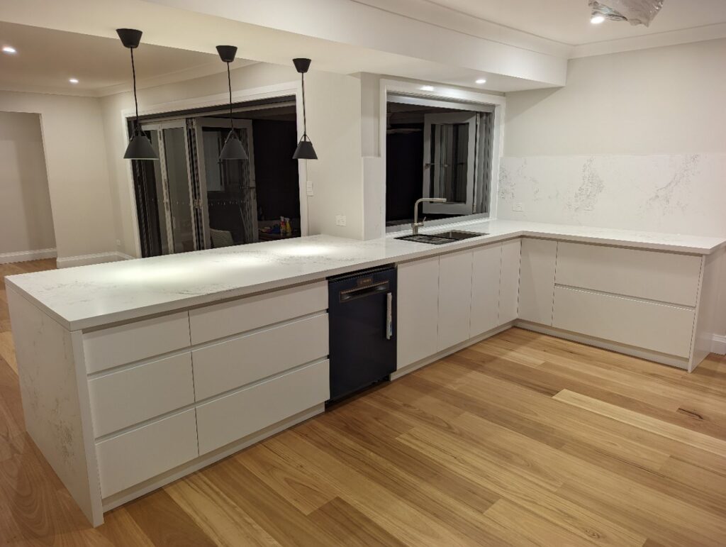 Modern kitchen with white cabinets and drawers, a marble countertop, built-in sink, black dishwasher, wooden floor, and three black pendant lights hanging above the counter. Large windows and sliding glass doors in the background.