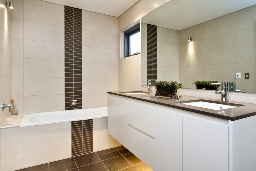 Modern bathroom with beige tiled walls, a built-in bathtub, a long white vanity with a dark countertop, two sinks, a large mirror, a small window, and a decorative plant on the counter.