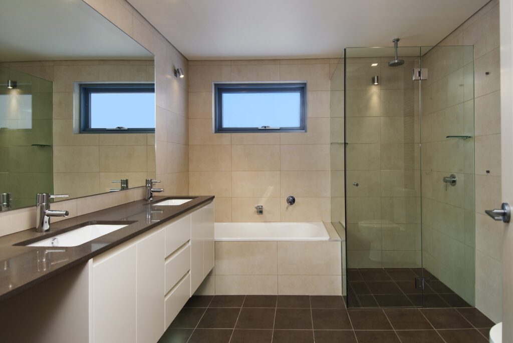 Modern bathroom with beige tiles, a glass-enclosed shower, a bathtub under a rectangular window, a double-sink vanity with a dark countertop, and large wall mirror.