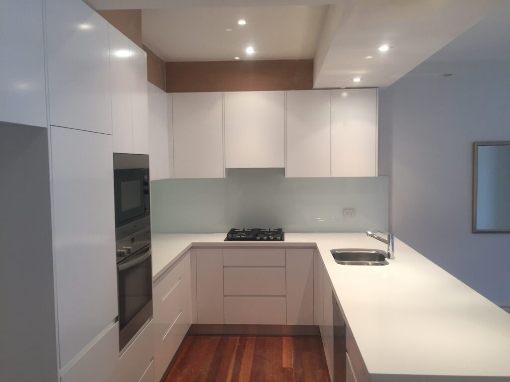Modern kitchen with white cabinets, stainless steel oven and microwave, a gas stovetop, minimalist faucet, glass backsplash, and wooden floor under bright ceiling lights.