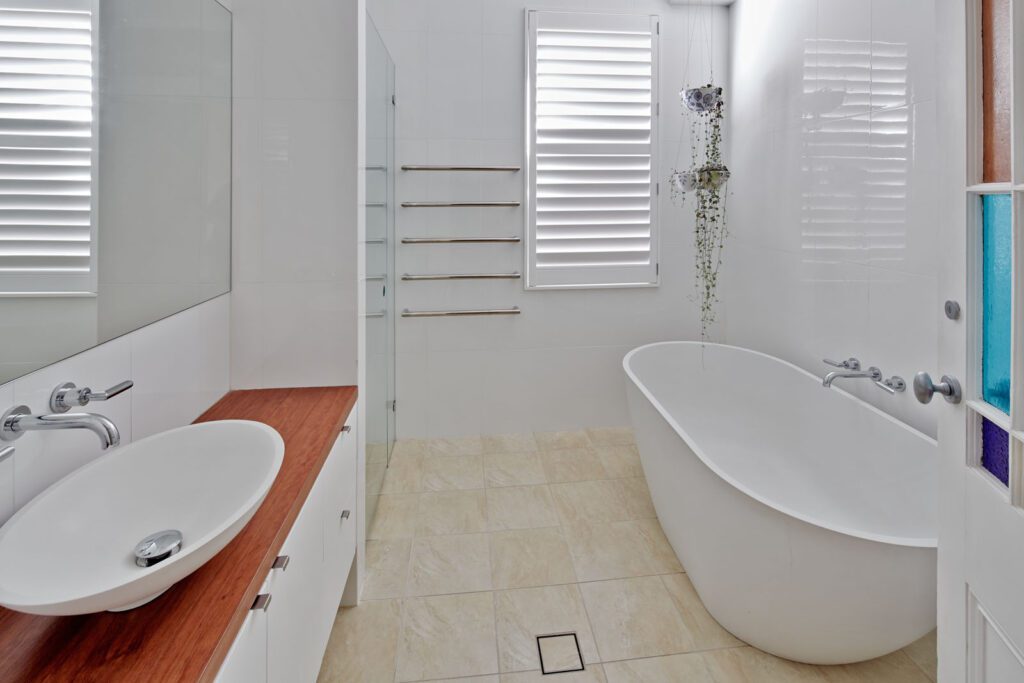 Modern bathroom with a freestanding white bathtub, vessel sink on a wooden countertop, wall-mounted towel racks, large window with white shutters, beige tiled floor, and a glass shower partition.