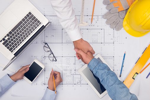 Two people shake hands over architectural blueprints, surrounded by a laptop, tablet, and hard hat—symbolizing collaboration in construction planning and the expertise of Building Services Sydney.